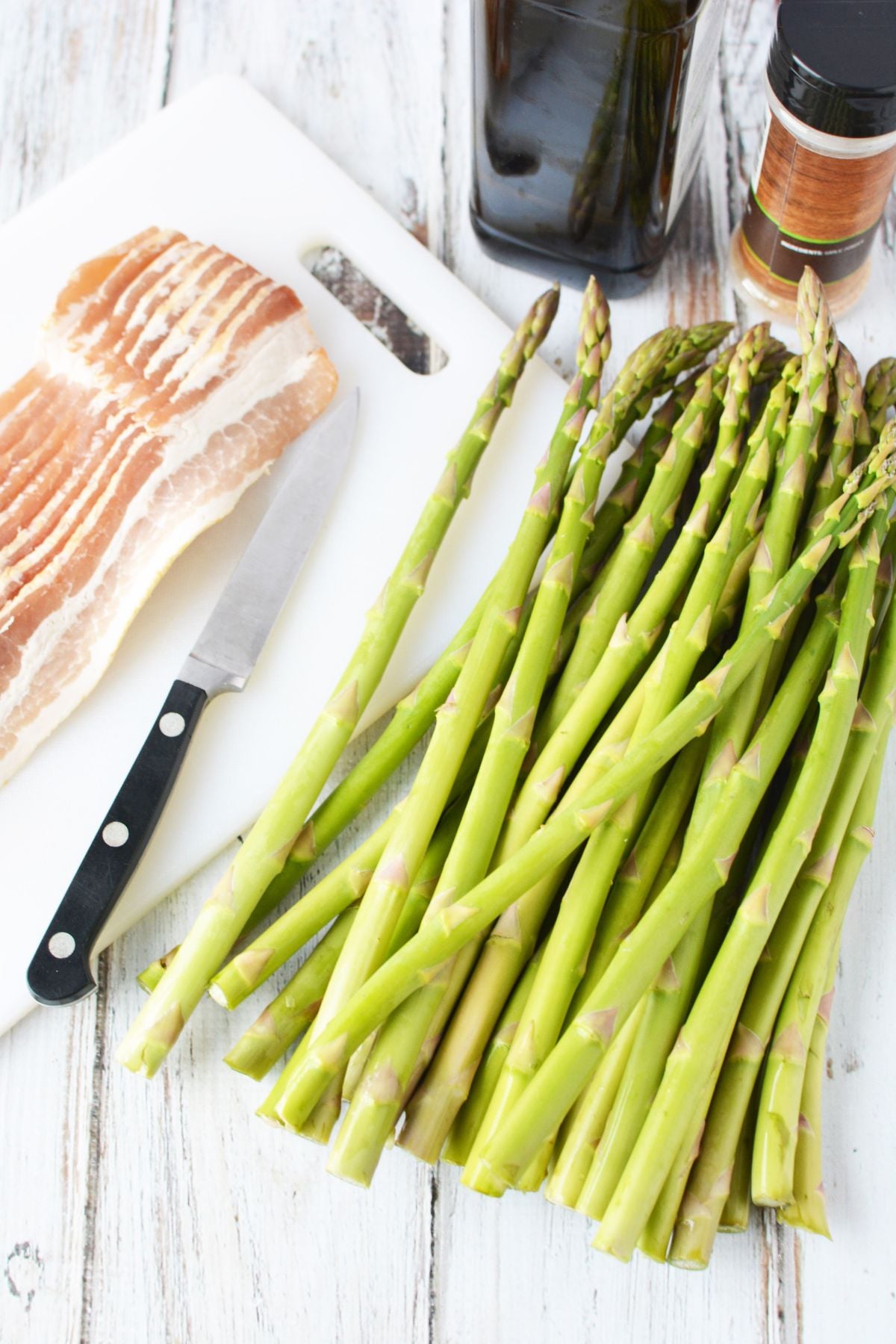 Fresh asparagus spears and raw bacon strips on white cutting board with knife and olive oil bottle