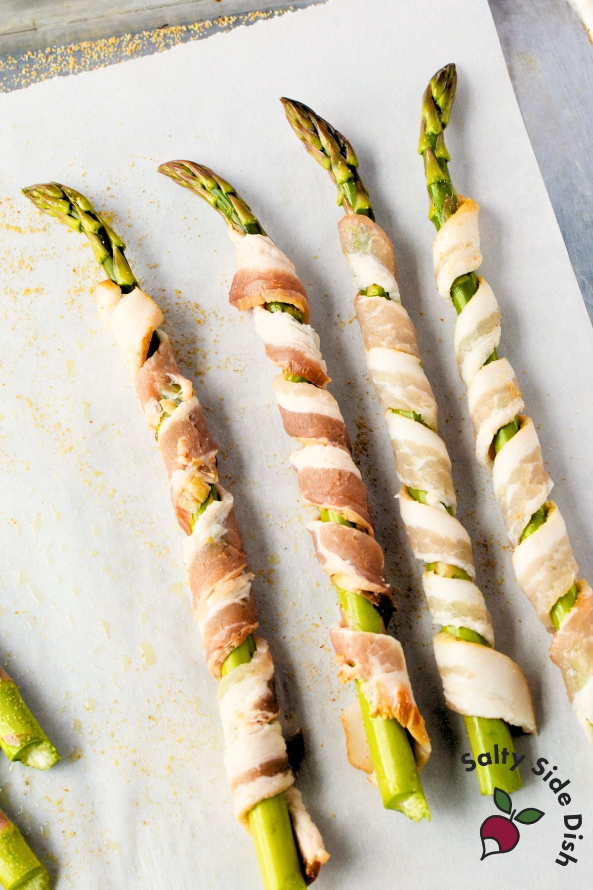 Bacon strips wrapped in spiral around fresh asparagus spears on parchment-lined baking sheet
