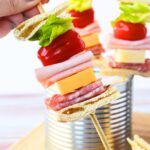 Close-up view of ingredients for sandwich on a stick, including sliced pita bread, chopped romaine lettuce, grape tomatoes, sliced salami, sliced ham, and cheese cubes, ready for assembly.