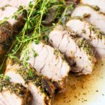 Close-up of sliced pork tenderloin with herb crust and fresh thyme on a white plate.