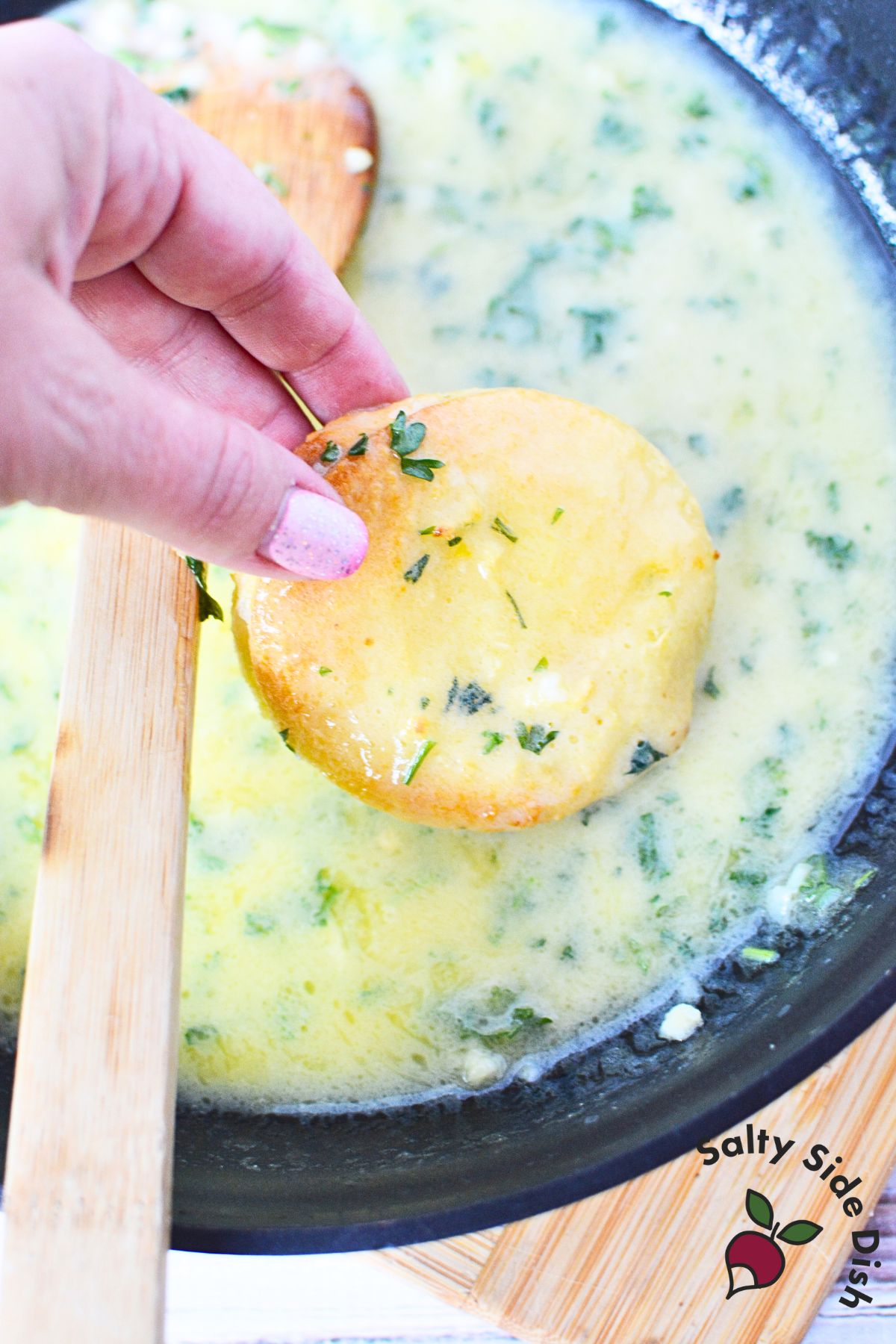 Hand dipping a crispy garlic bread round into a skillet of warm garlic butter sauce with herbs.
