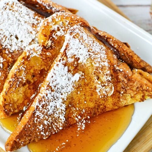 Plate of golden brioche French toast served with powdered sugar and maple syrup, showing crispy edges and soft center.