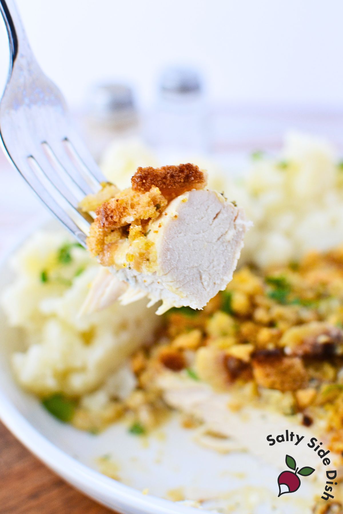 Fork holding tender bite of slow cooked chicken breast with stuffing topping and mashed potatoes in background