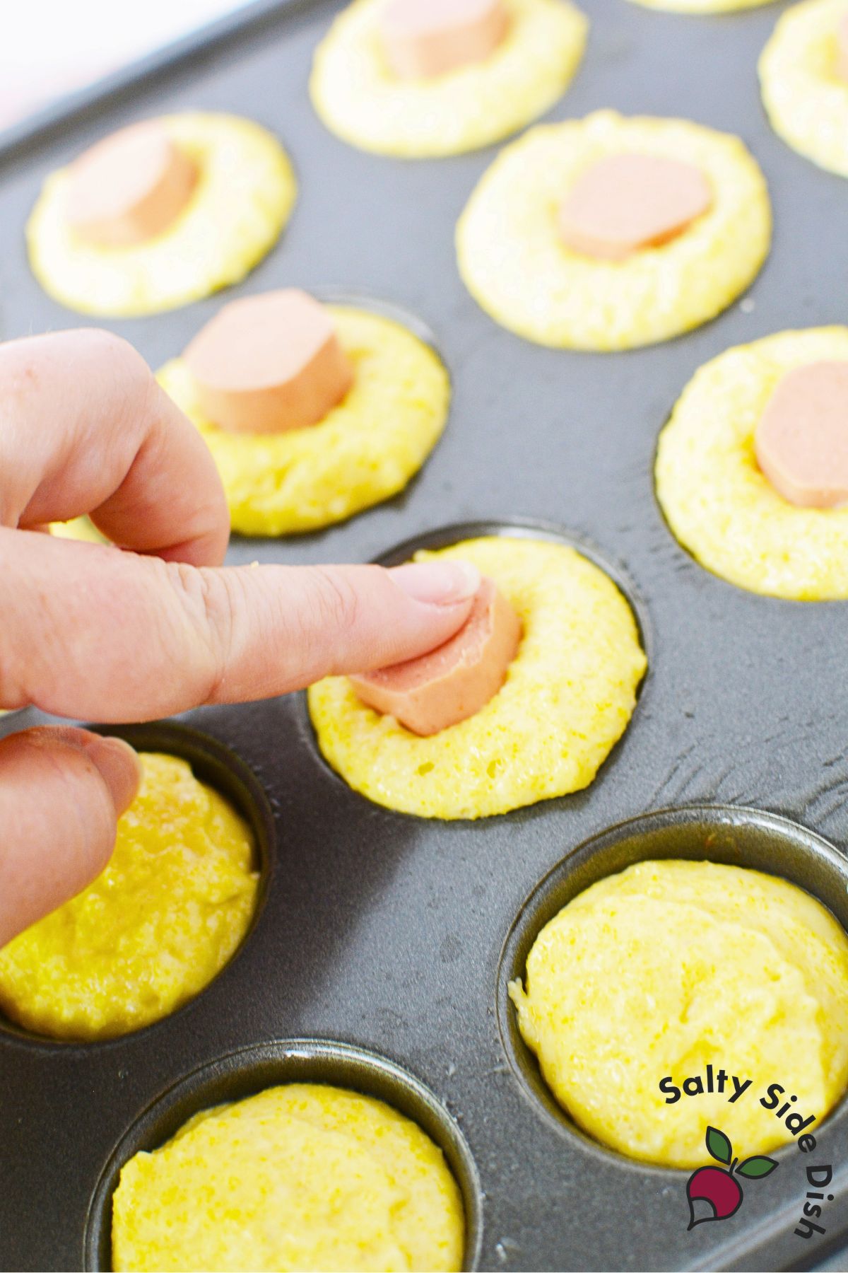 Close-up of a hand pressing a cut hot dog piece into the center of Jiffy corn muffin batter in a mini muffin tin