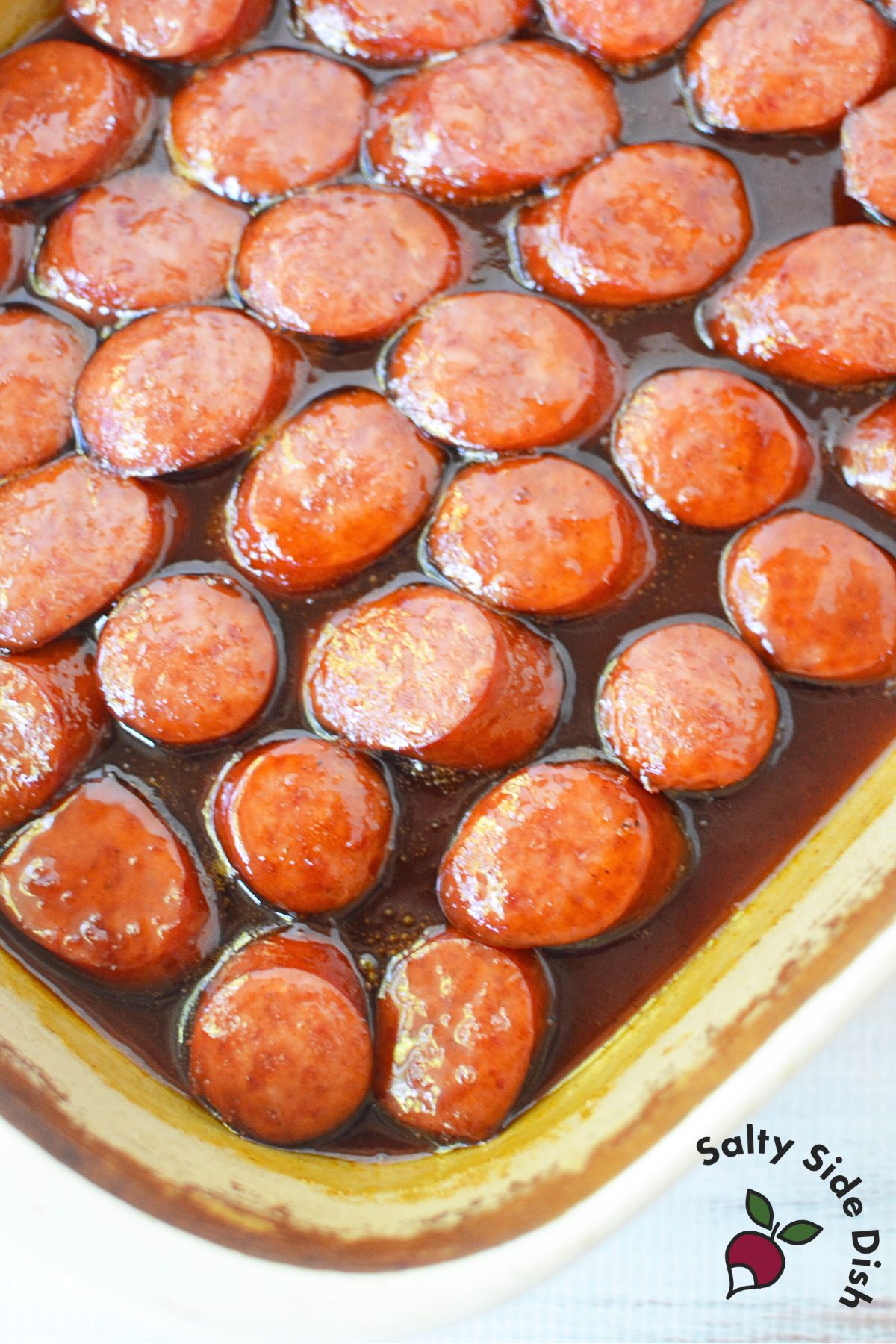 Sliced smoked sausage layered in baking dish with brown sugar and BBQ sauce being mixed for easy oven appetizer