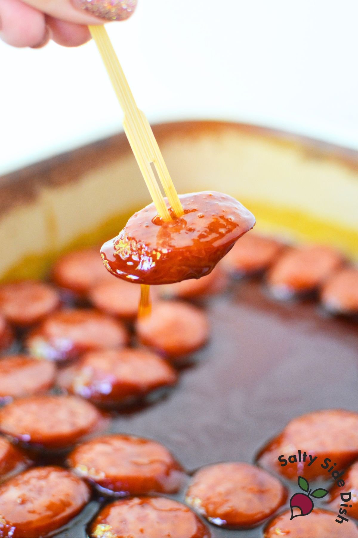 Single sausage slice dipped in BBQ glaze lifted on appetizer toothpick for serving