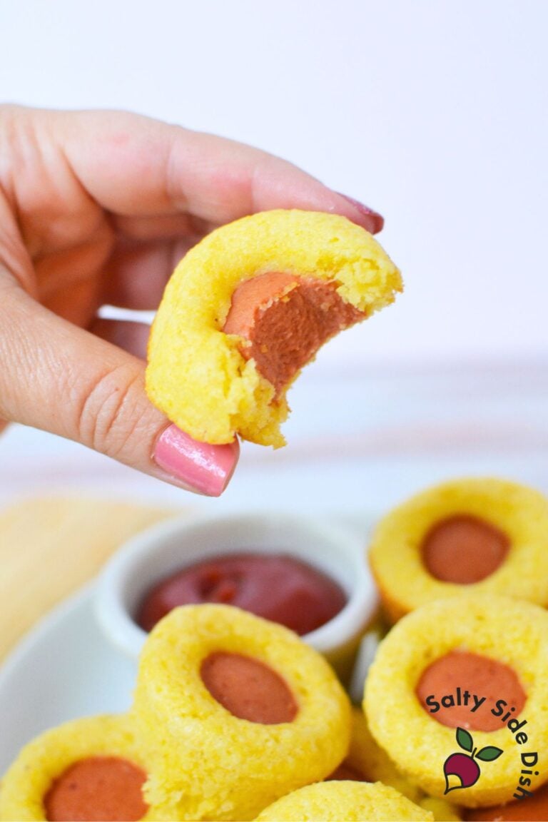 A hand holding one whole baked mini corn dog muffin with a visible hot dog center and golden edges