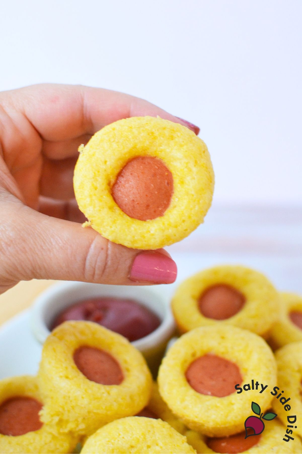 A hand holding a mini corn dog muffin with a bite taken out to show the hot dog center and fluffy cornbread texture