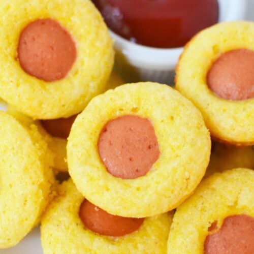 Baked mini corn dog muffins stacked on a white plate next to a ramekin of ketchup for dipping