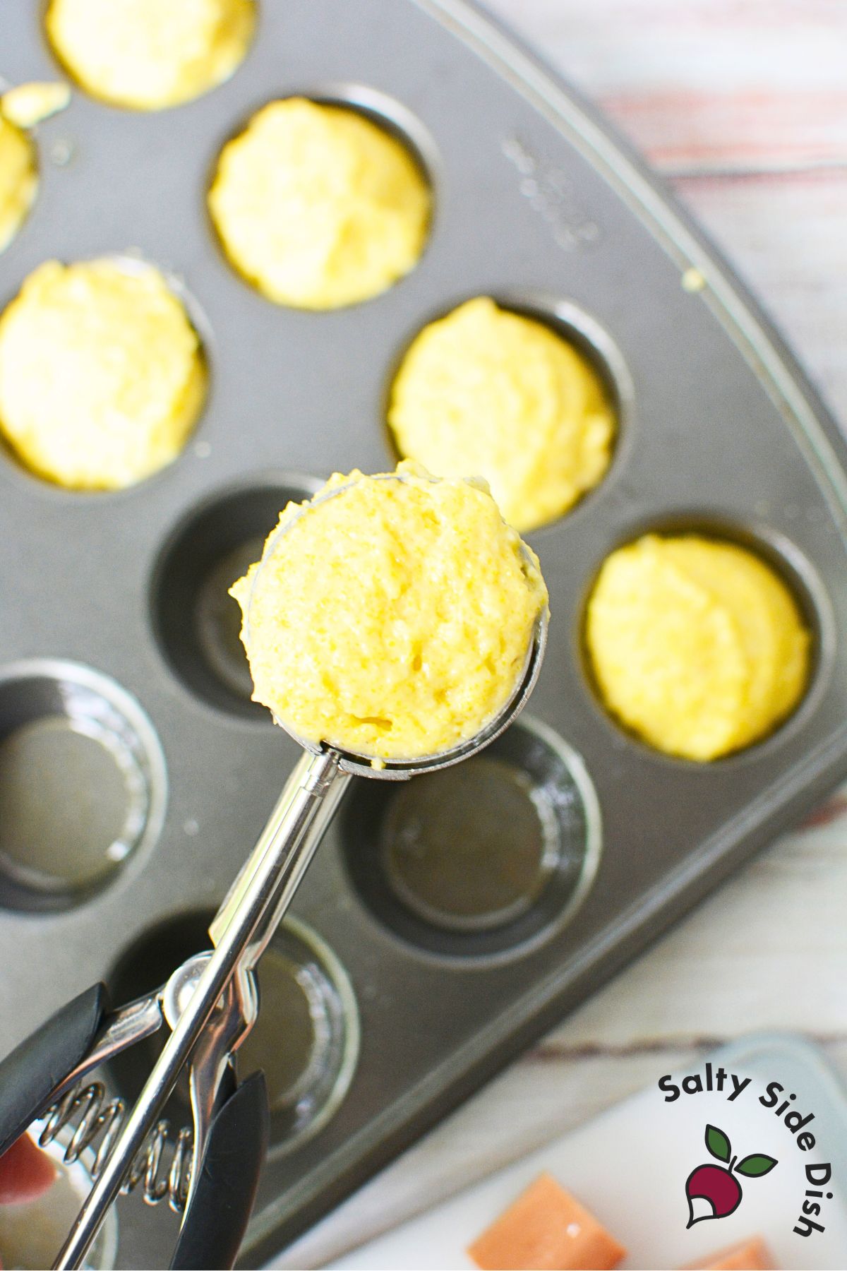 A tablespoon cookie scoop filled with yellow cornbread batter held over a greased mini muffin tin for corn dog muffins