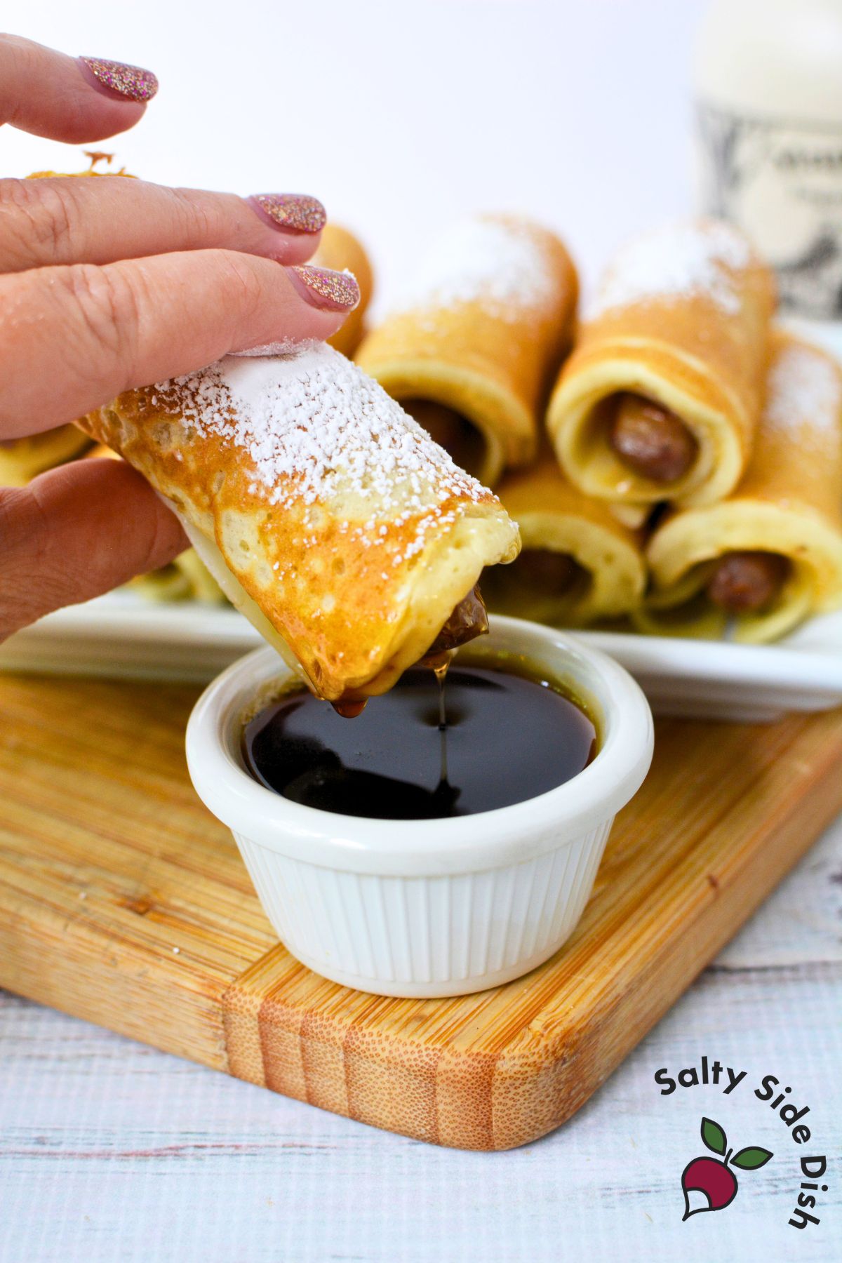 Hand holding a powdered sugar-topped pancake sausage roll and dipping it into a cup of maple syrup.