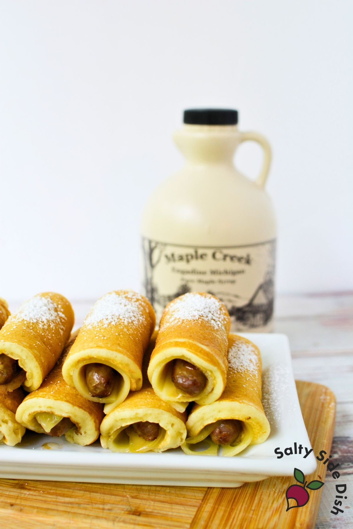 A plate of pancake-wrapped sausages in front of a bottle of Maple Creek pure maple syrup.