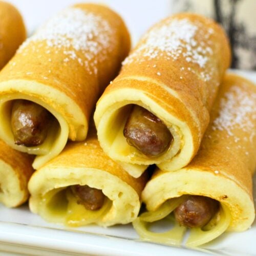 Golden brown pancake-wrapped sausages dusted with powdered sugar on a white plate, served with syrup.