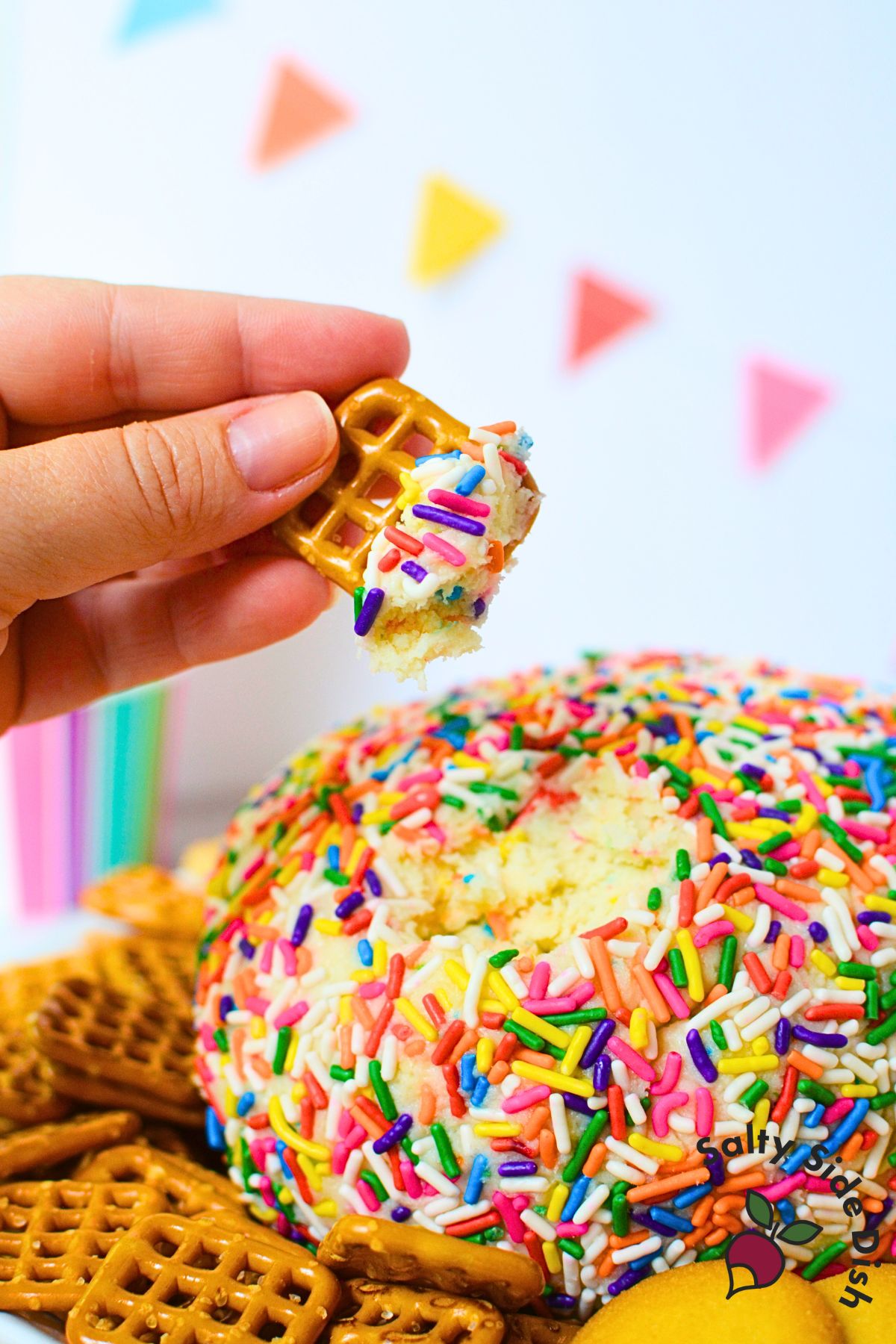 Close up of a sweet birthday cake cheese ball coated in rainbow sprinkles, surrounded by cookies and crackers.