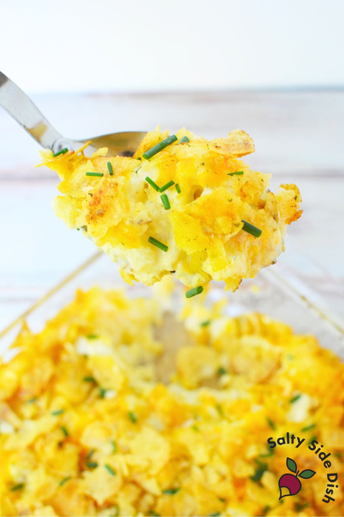 Spoonful of creamy sour cream and onion potato casserole with cheddar and chip topping being lifted from baking dish.