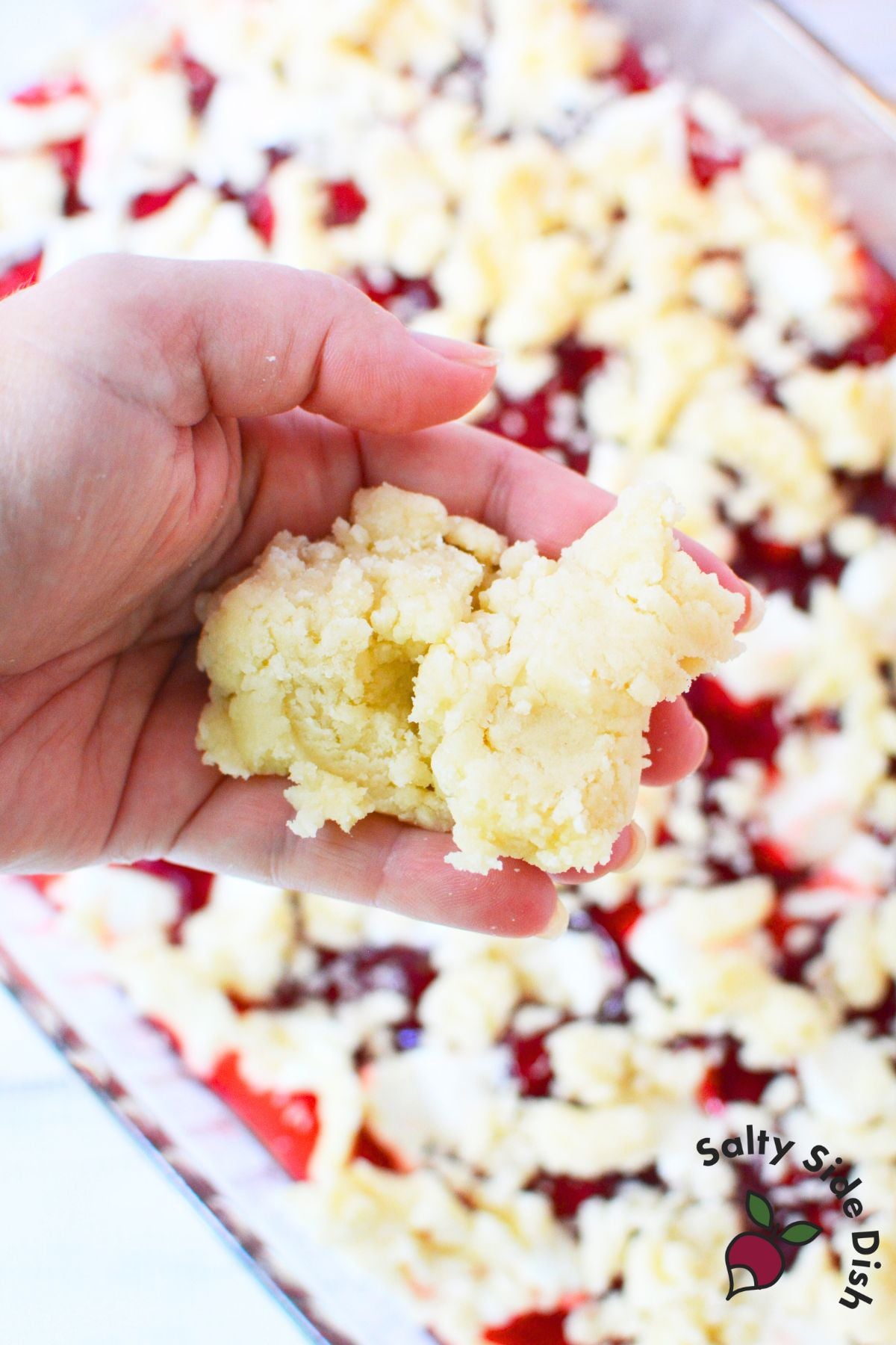 Hand holding buttery cake mix topping before adding it over strawberry and cream cheese layers.