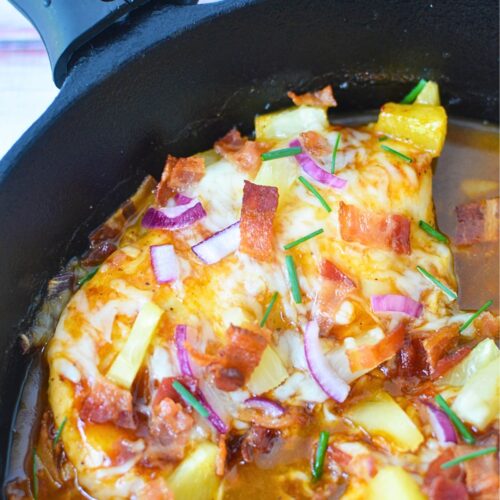 Overhead shot of unbaked Hawaiian chicken in a cast iron skillet with pineapple, bacon, red onions, and cheese.