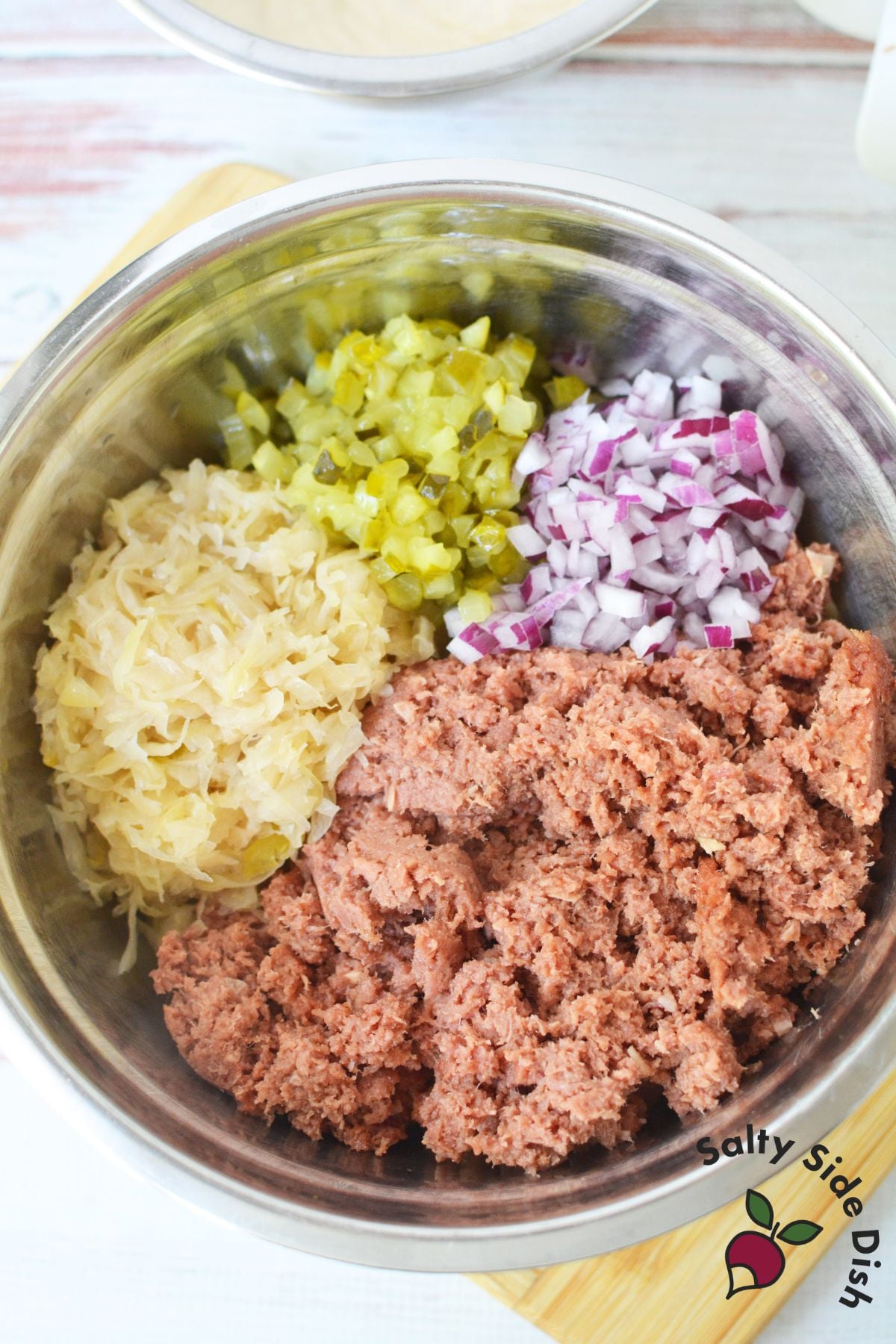 Bowl with canned corned beef, sauerkraut, diced pickles, and red onion ready to mix into Reuben sandwich pasta salad.