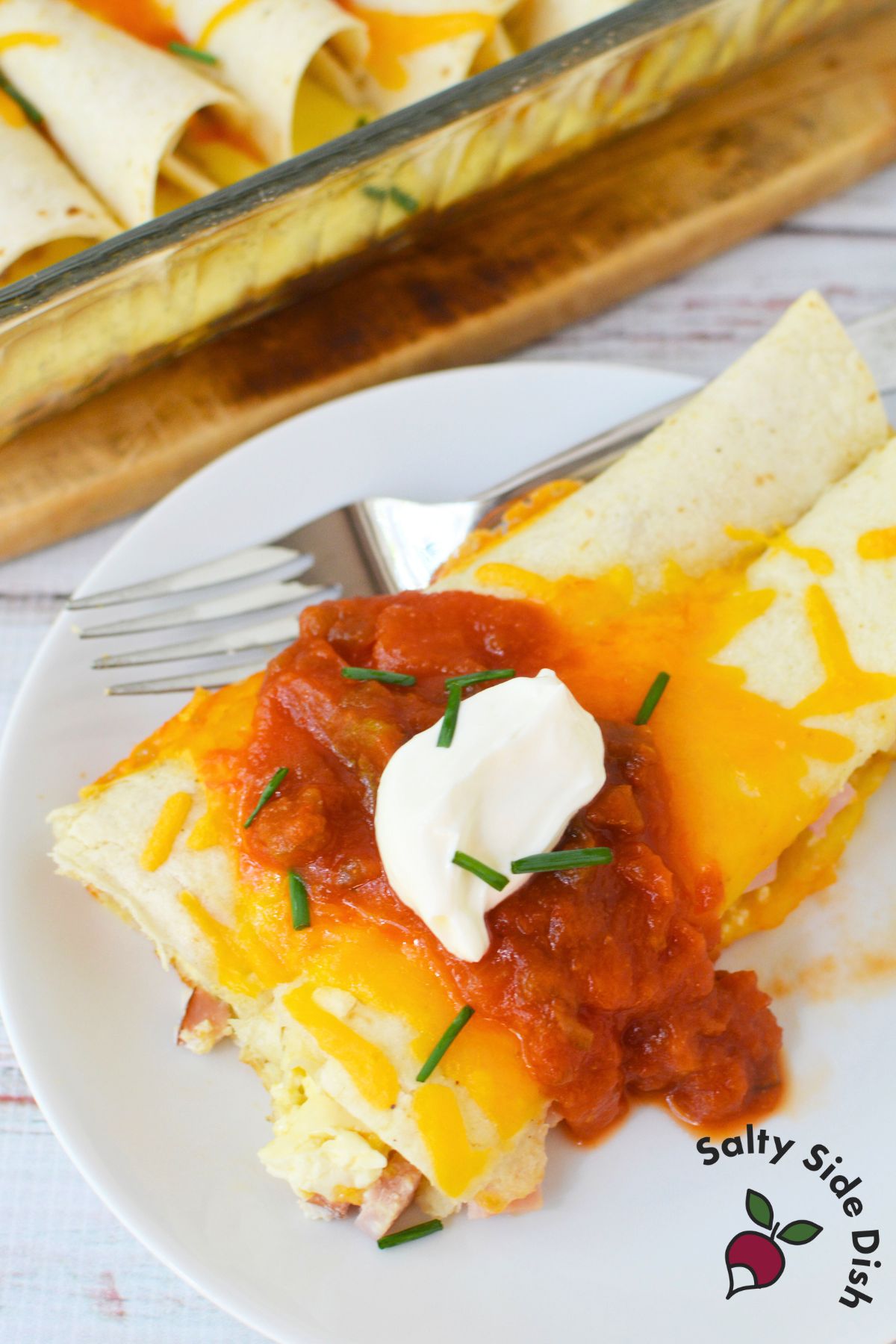 A serving of cheesy ham and egg enchilada topped with sour cream and salsa on a white plate with fork.