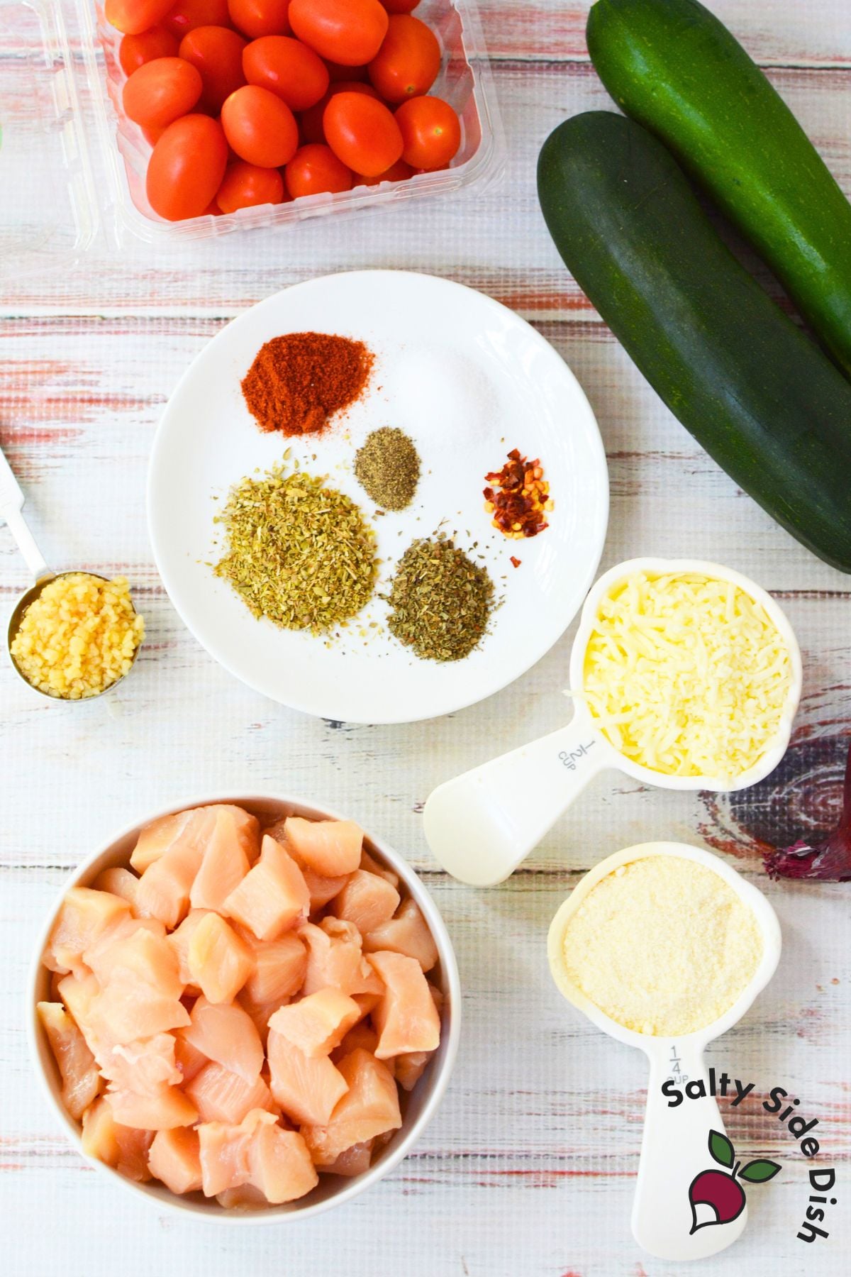 Raw chicken, zucchini, tomatoes, seasonings, and cheese displayed for Mediterranean chicken bake.