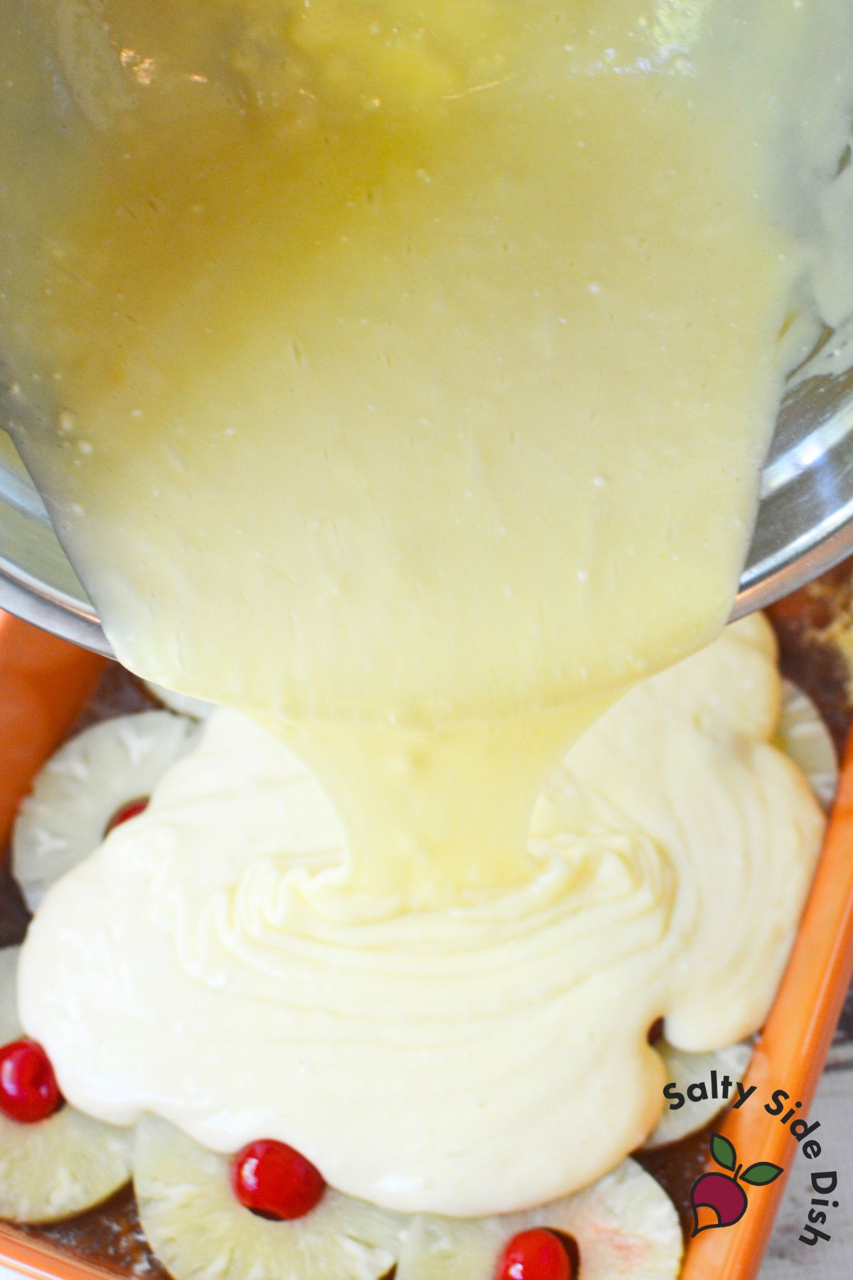 White cake batter being poured over pineapple rings and cherries in a baking dish for pineapple upside down cake.