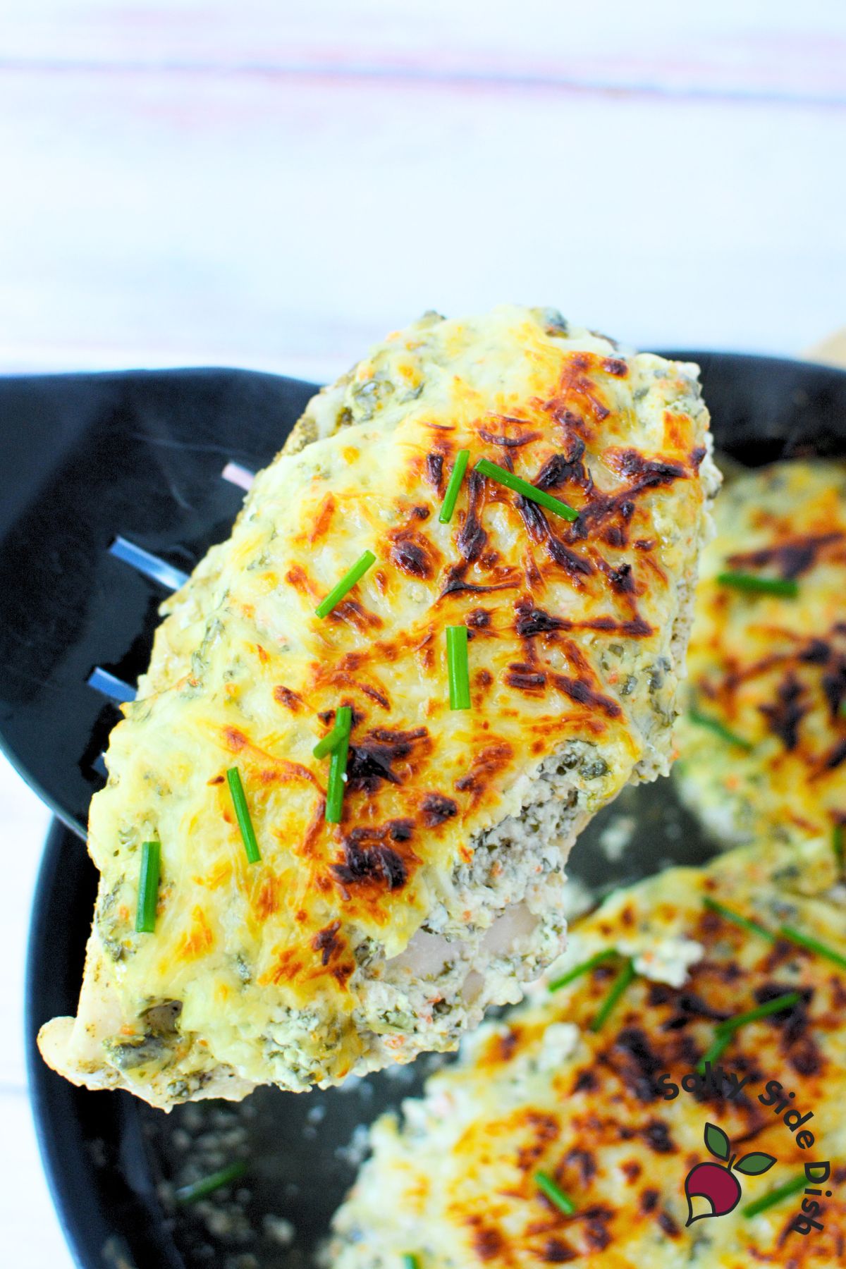 Slice of baked spinach dip chicken on a white plate with golden cheese and chives.