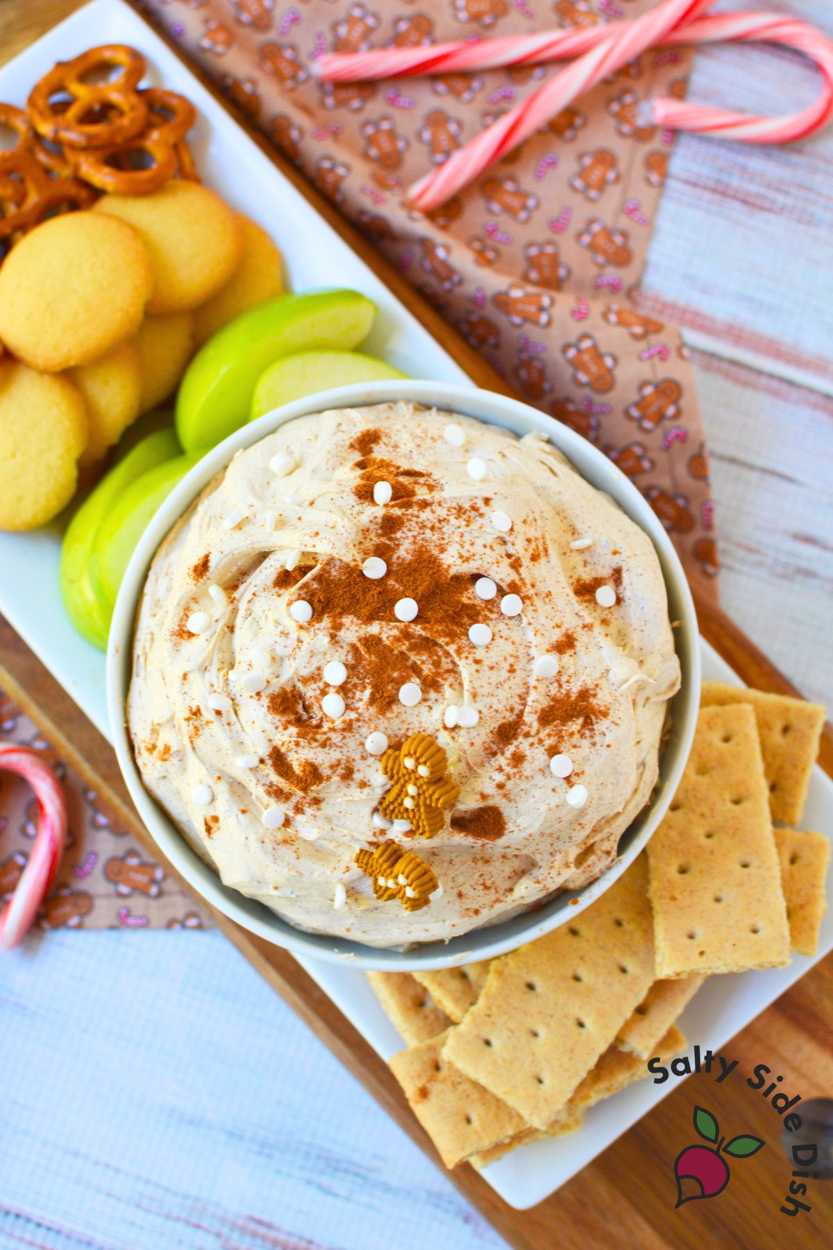 Bowl of gingerbread dip with cinnamon, white sprinkles, and gingerbread men decorations surrounded by pretzels, graham crackers, and apple slices.