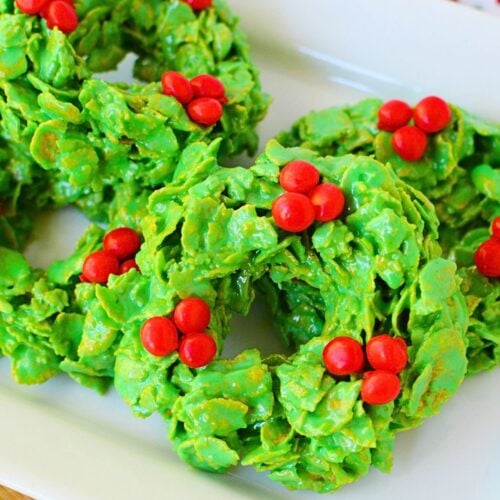 Holiday cornflake wreath cookies stacked on a platter, decorated with red candies.