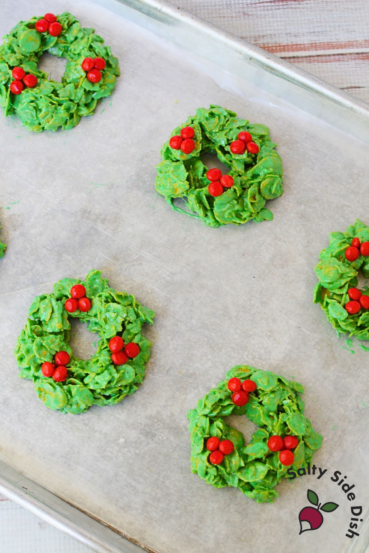 Green cornflake wreath cookies with red candies setting on a wax paper lined baking sheet.
