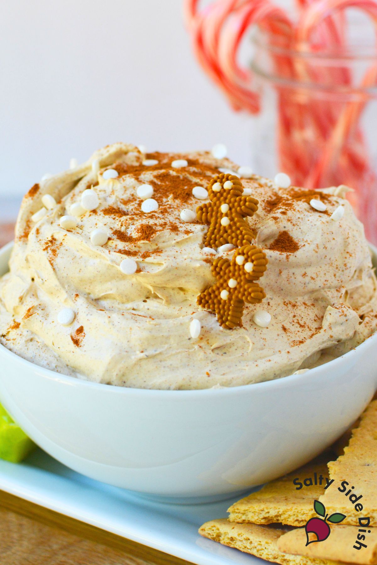 Festive gingerbread dip in a bowl decorated with sprinkles and cinnamon surrounded by crackers and apple slices.