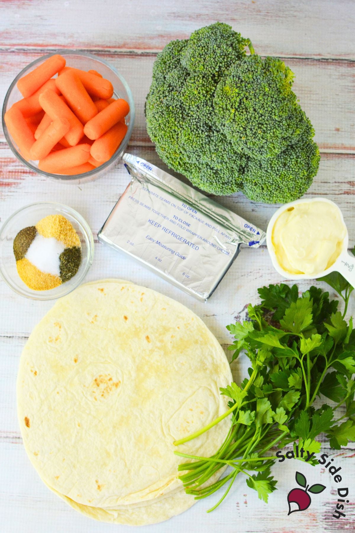 Ingredients for veggie pinwheels laid out on a table including tortillas, cream cheese, mayonnaise, carrots, broccoli, parsley, and spices.