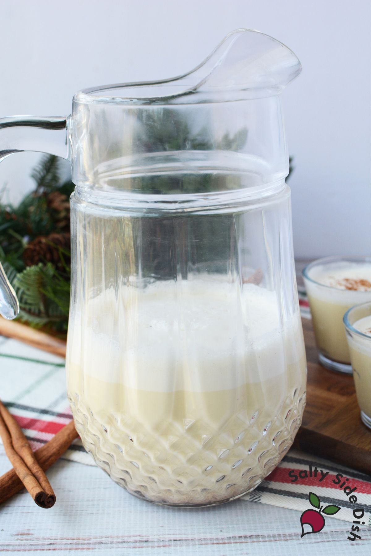 Glass pitcher of homemade spiked eggnog on a holiday table with cinnamon sticks and Christmas greenery