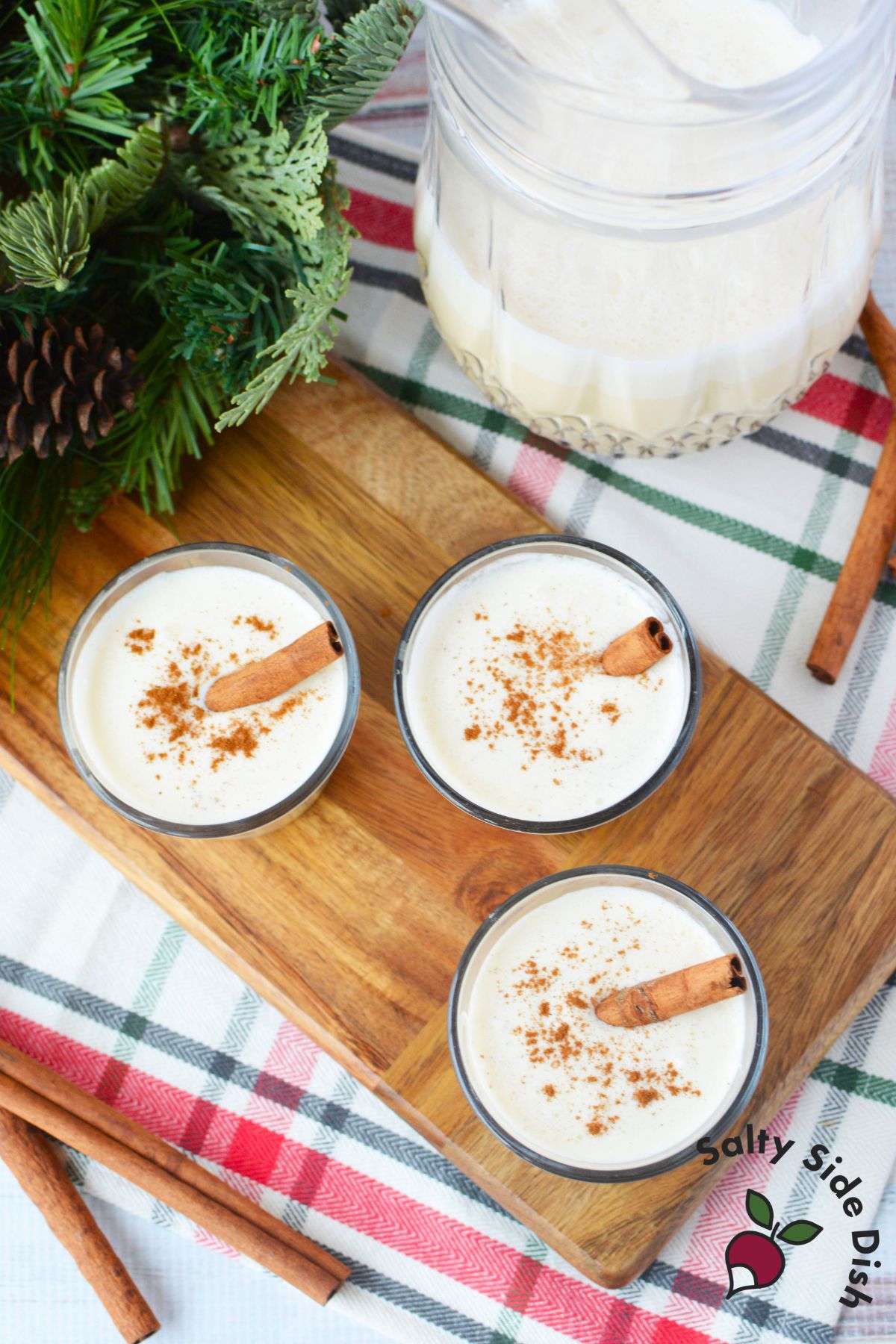 Small glasses of creamy spiked eggnog topped with ground cinnamon on a wooden serving board