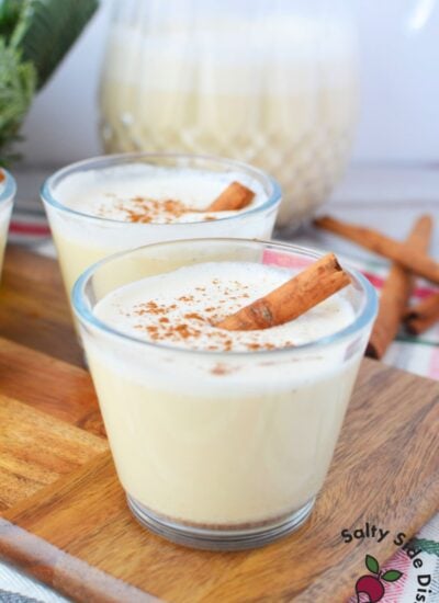 Two glasses of homemade Christmas spiked eggnog with cinnamon sticks in front of glass pitcher