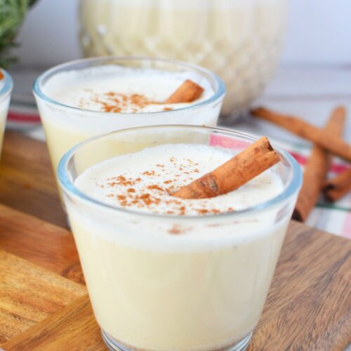 Two glasses of homemade Christmas spiked eggnog with cinnamon sticks in front of glass pitcher