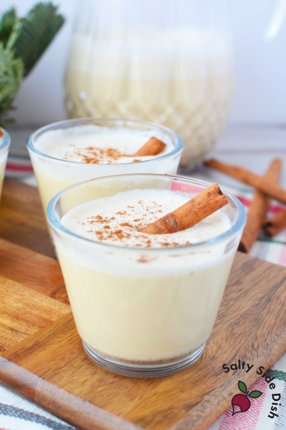 Two glasses of homemade Christmas spiked eggnog with cinnamon sticks in front of glass pitcher
