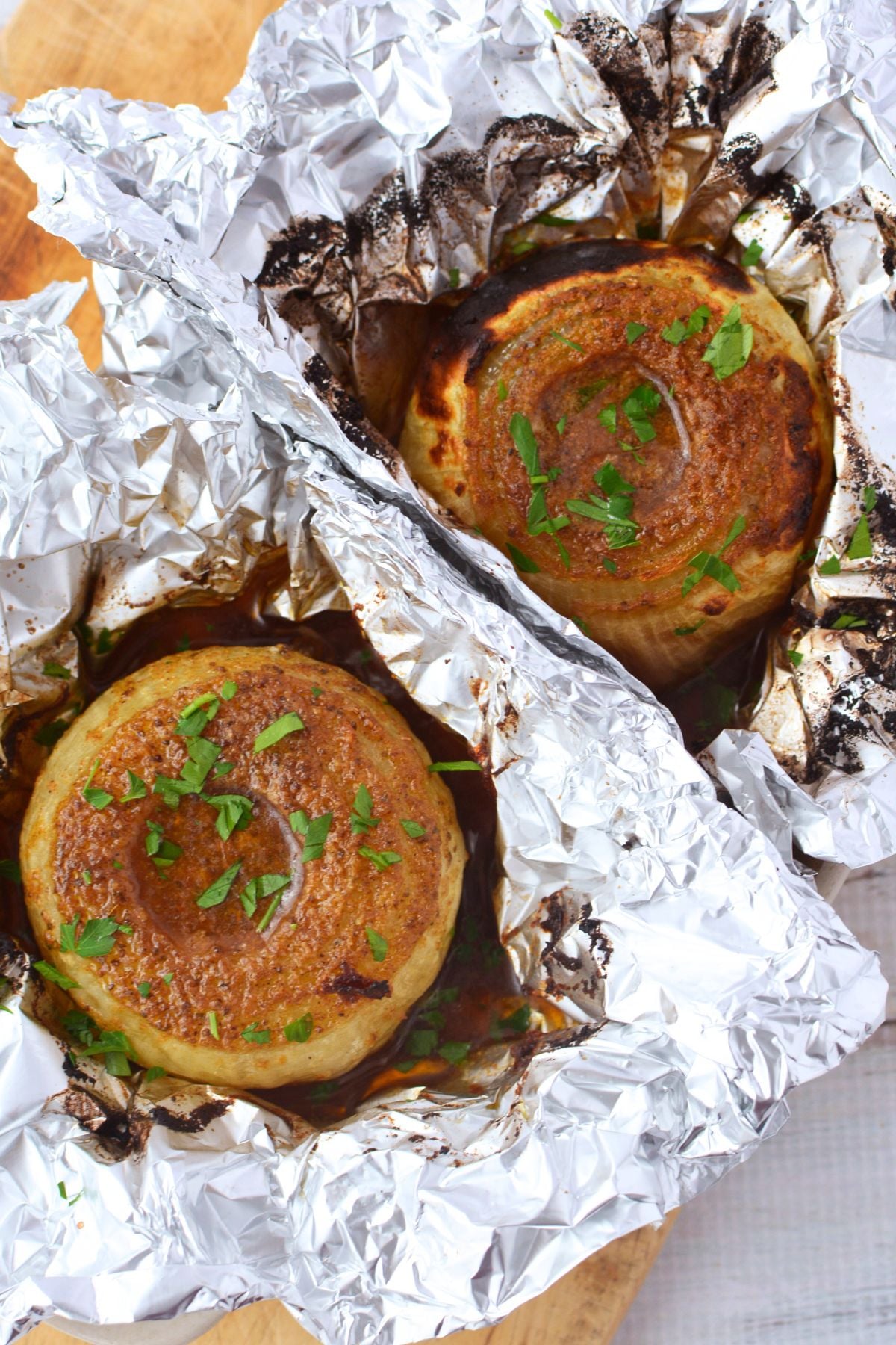 Two whole baked onions with golden caramelized Old Bay tops garnished with fresh parsley sitting in foil in a baking dish