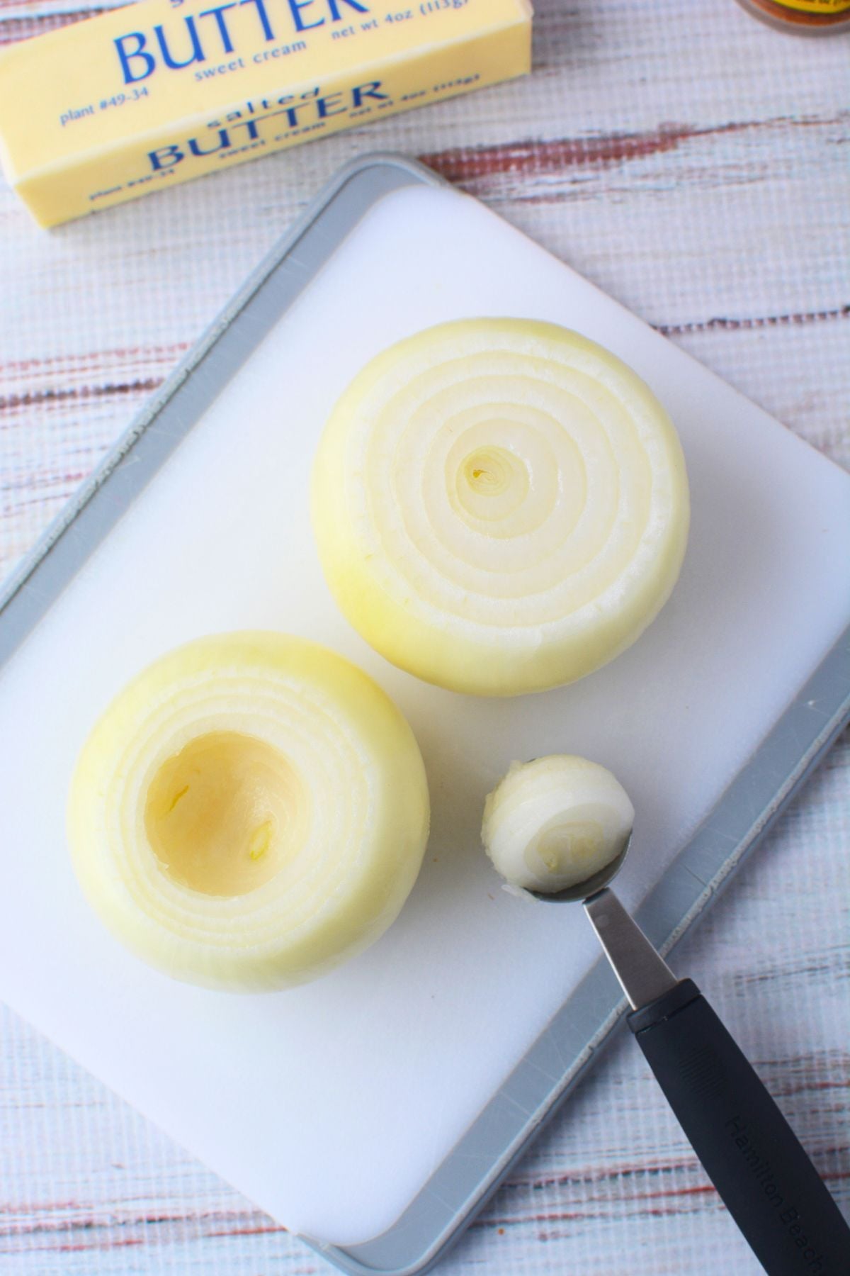Two peeled sweet onions on a white cutting board, one hollowed out with a melon baller and one whole, with a stick of butter nearby