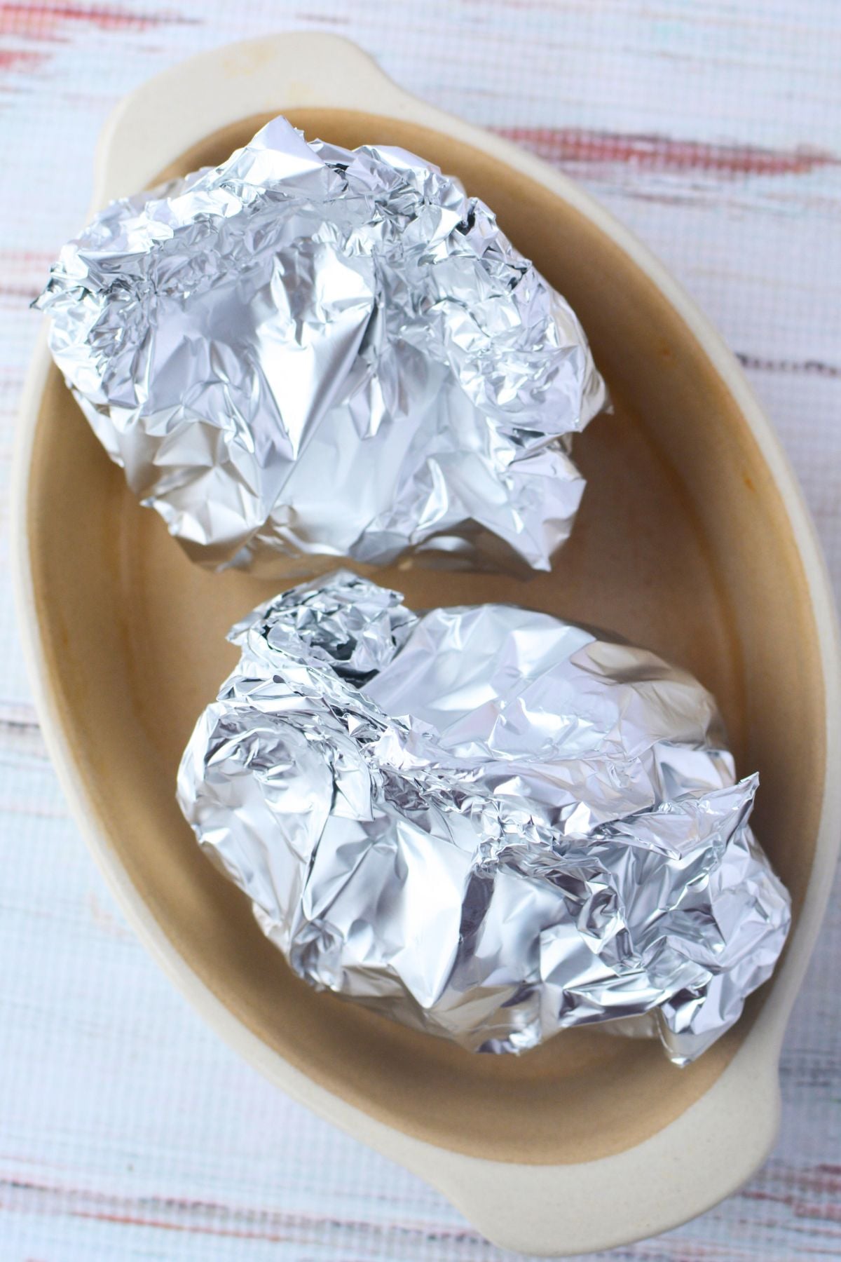Two foil-wrapped onions placed upright in a shallow ceramic baking dish ready for the oven
