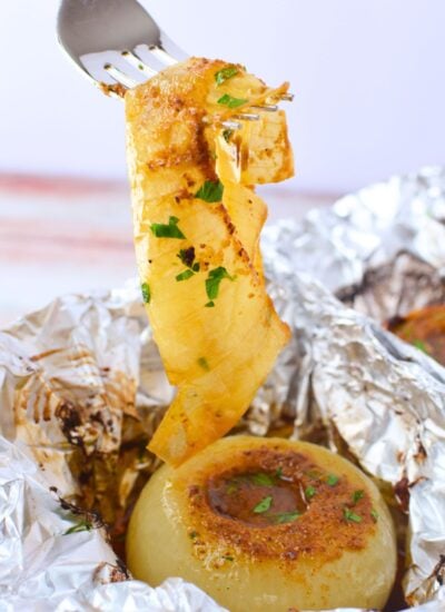 A fork lifting soft caramelized onion layers from a baked onion boil sitting in foil with buttery Old Bay juices