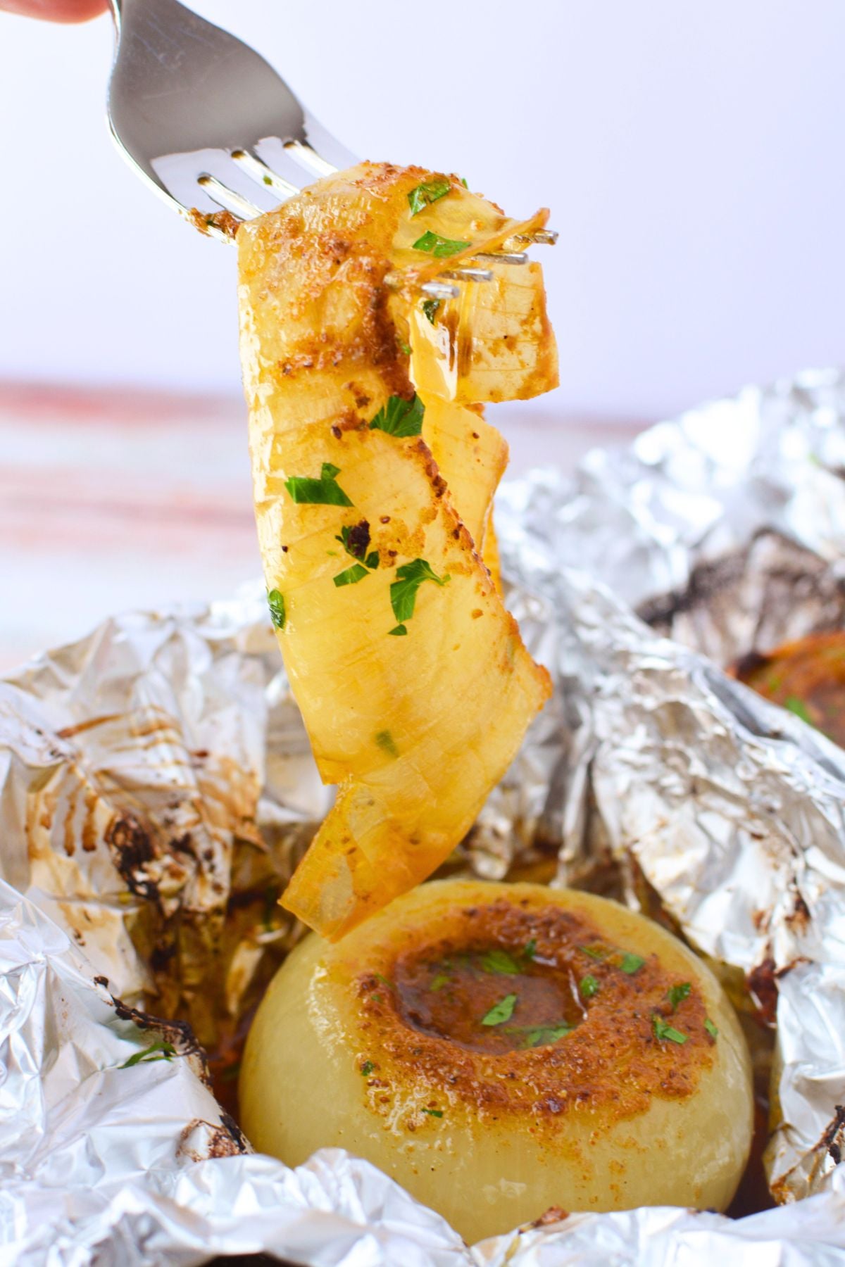A fork lifting soft caramelized onion layers from a baked onion boil sitting in foil with buttery Old Bay juices