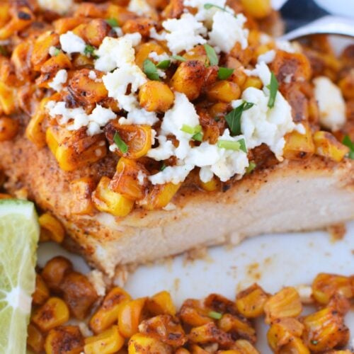 Close up of sliced baked chicken breast topped with charred elote corn, queso fresco, and a lime wedge