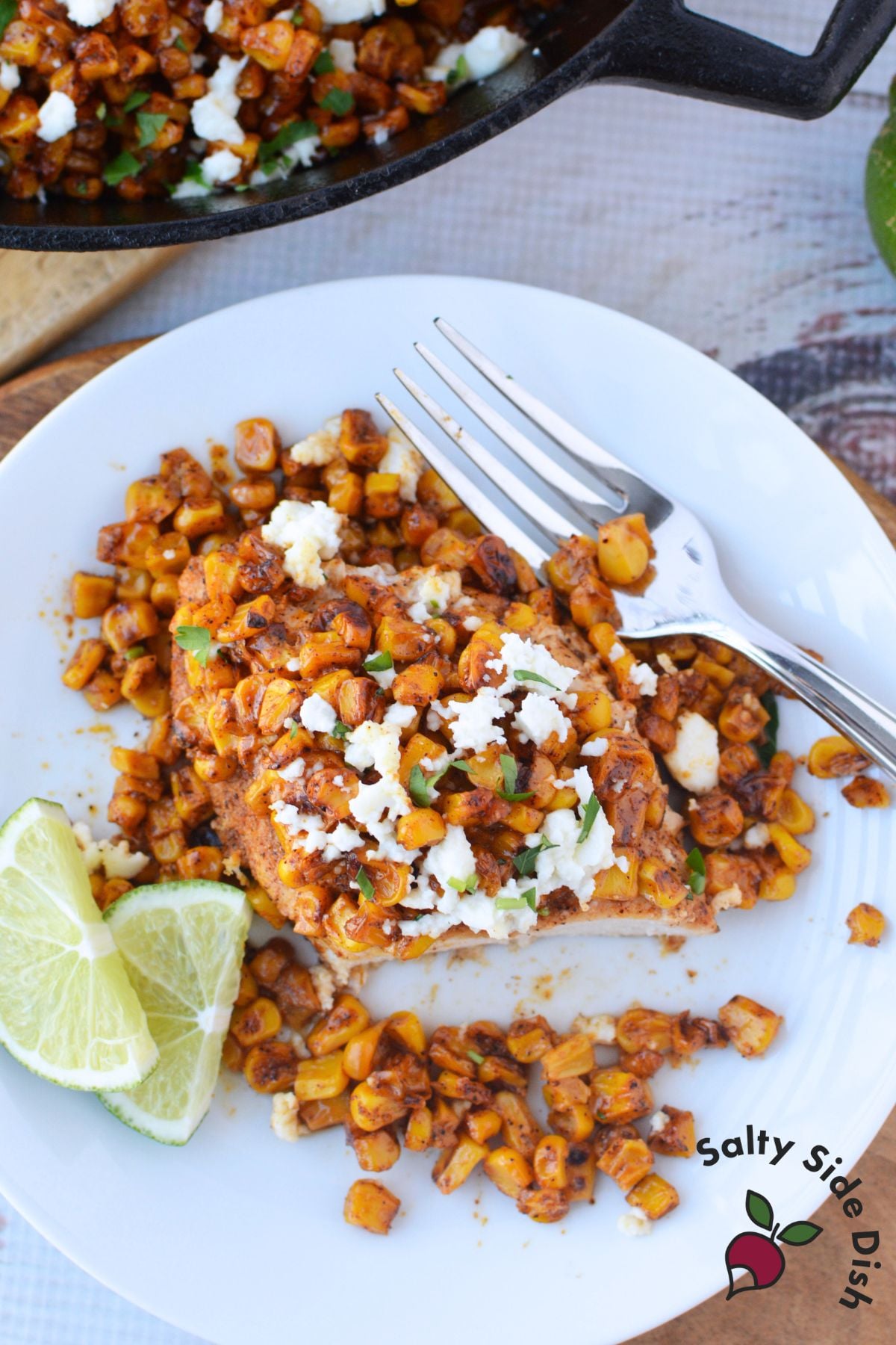 Plated serving of Mexican street corn chicken bake with charred corn, queso fresco crumbles, and lime wedges with the cast iron skillet in the background