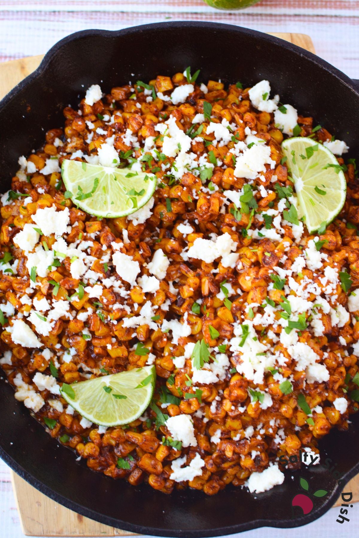Mexican street corn chicken bake in a cast iron skillet topped with crumbled queso fresco, cilantro, and lime wedges