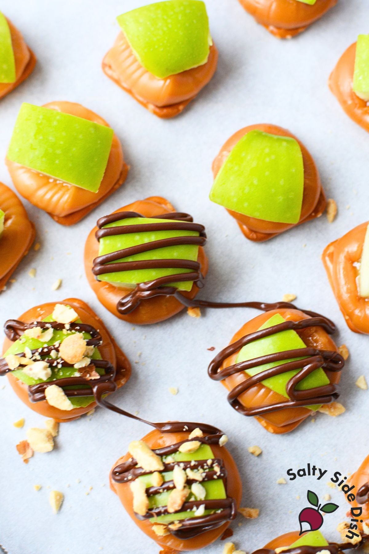 Caramel apple pretzel bites in various stages with chocolate being drizzled over granny smith apple pieces and chopped peanuts on parchment paper