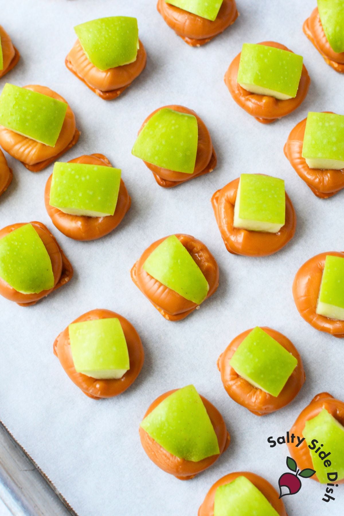 Granny smith apple pieces pressed into softened caramel on square pretzel snaps on a parchment lined baking sheet