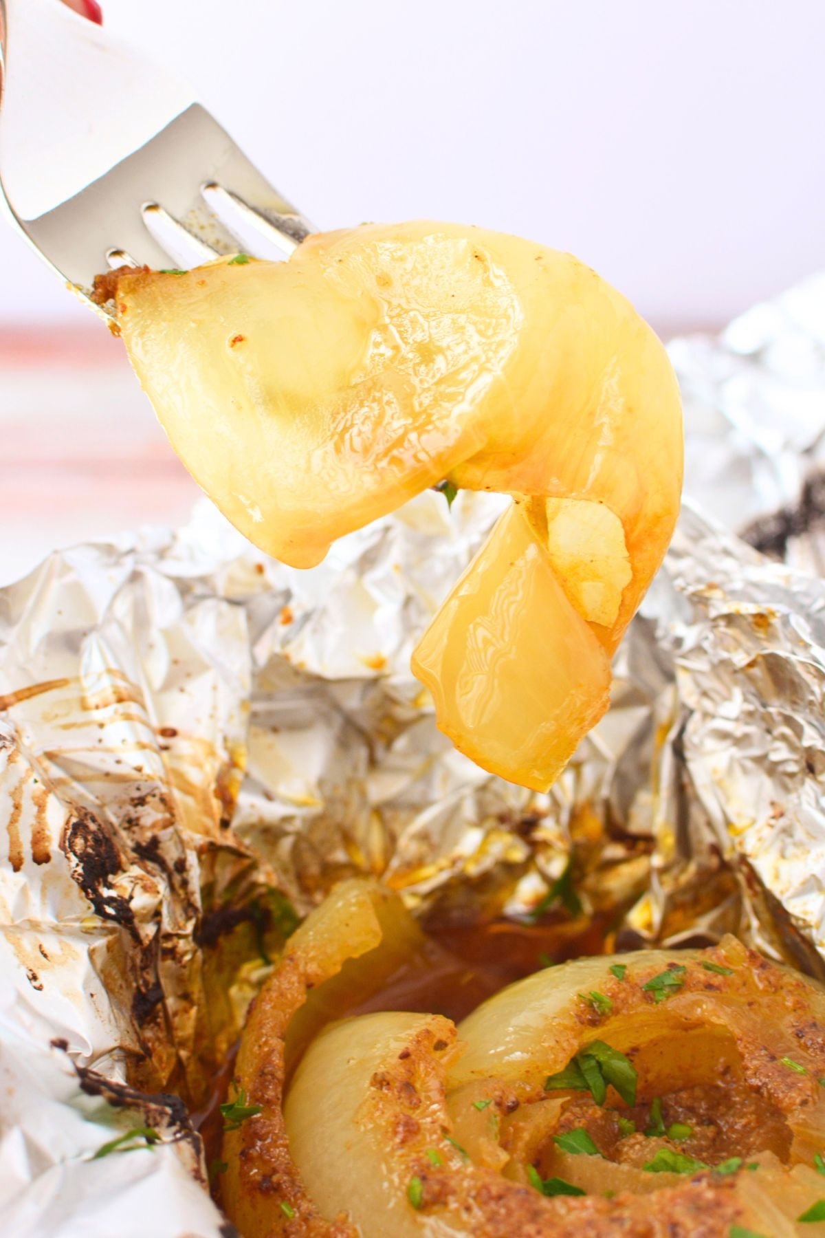 A fork holding soft translucent caramelized onion layers from a baked onion boil with the finished onion and buttery juices in foil below