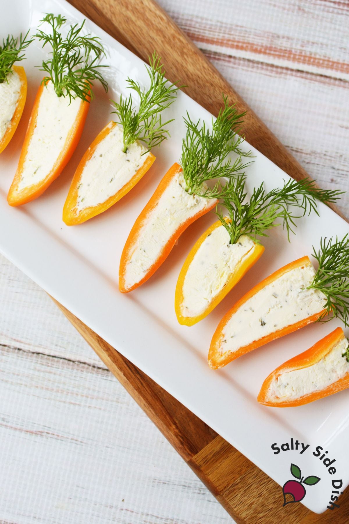 Hand holding a finished stuffed mini pepper carrot with Boursin cheese and fresh dill with platter of carrots in the background