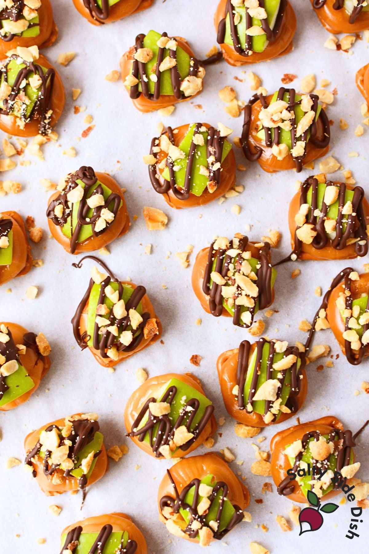 Overhead view of finished caramel apple pretzel bites with chocolate drizzle and chopped peanuts scattered across parchment paper