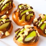 Hand holding a single caramel apple pretzel bite with chocolate drizzle and peanuts with platter of bites in the background